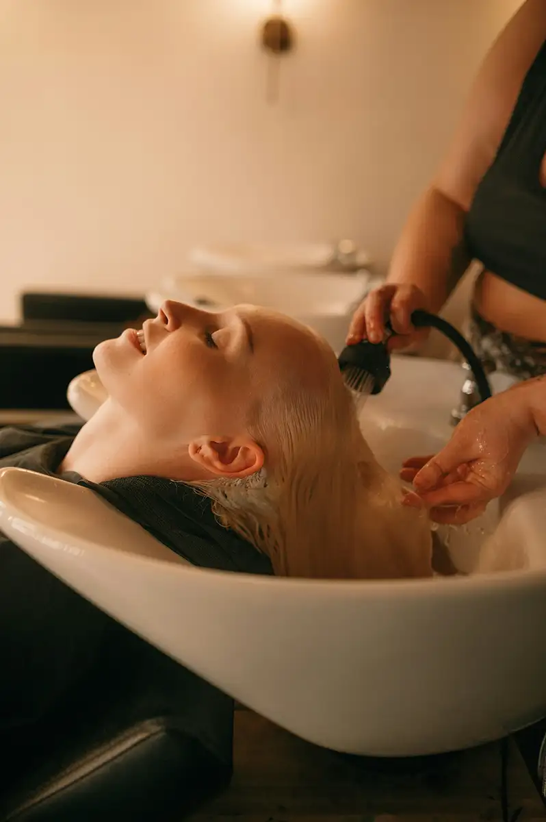 En person der ligger tilbagebøjet ved en salonvask, mens en stylist vasker håret med en håndholdt bruser.